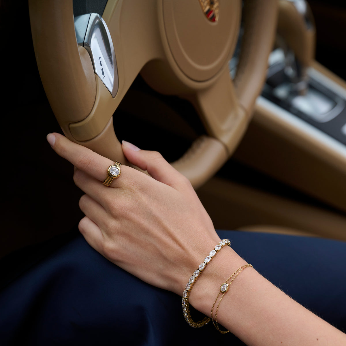 Close-up lifestyle shot of a woman's hand on a car steering wheel, wearing a gold ring with a round clear stone and ridged band, paired with a tennis bracelet and a fine chain bracelet.
Photo lifestyle en gros plan de la main d'une femme sur un volant, portant une bague dorée sertie d'une pierre transparente ronde à anneau strié, agencée avec un bracelet tennis et un bracelet à chaîne fine.